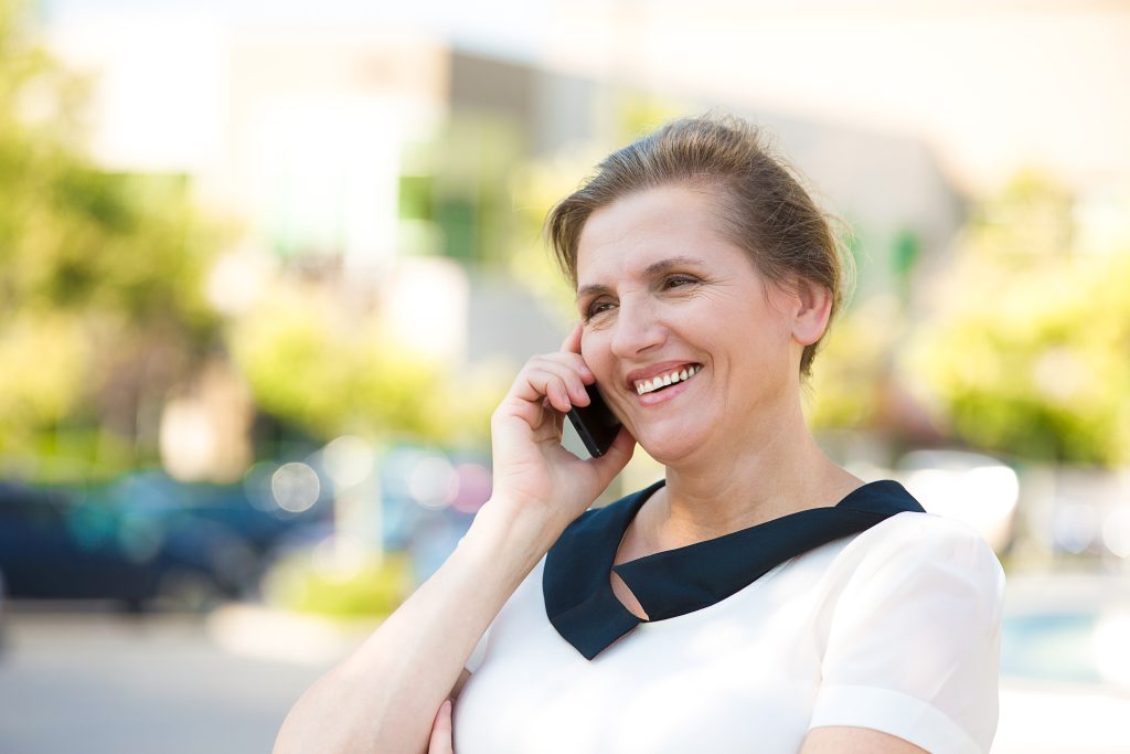 With a captive audience, Message On Hold is the perfect time to suggest complementary products or services. - Woman smiling while holding phone to her ear showing an existing customer listening to a message on hold